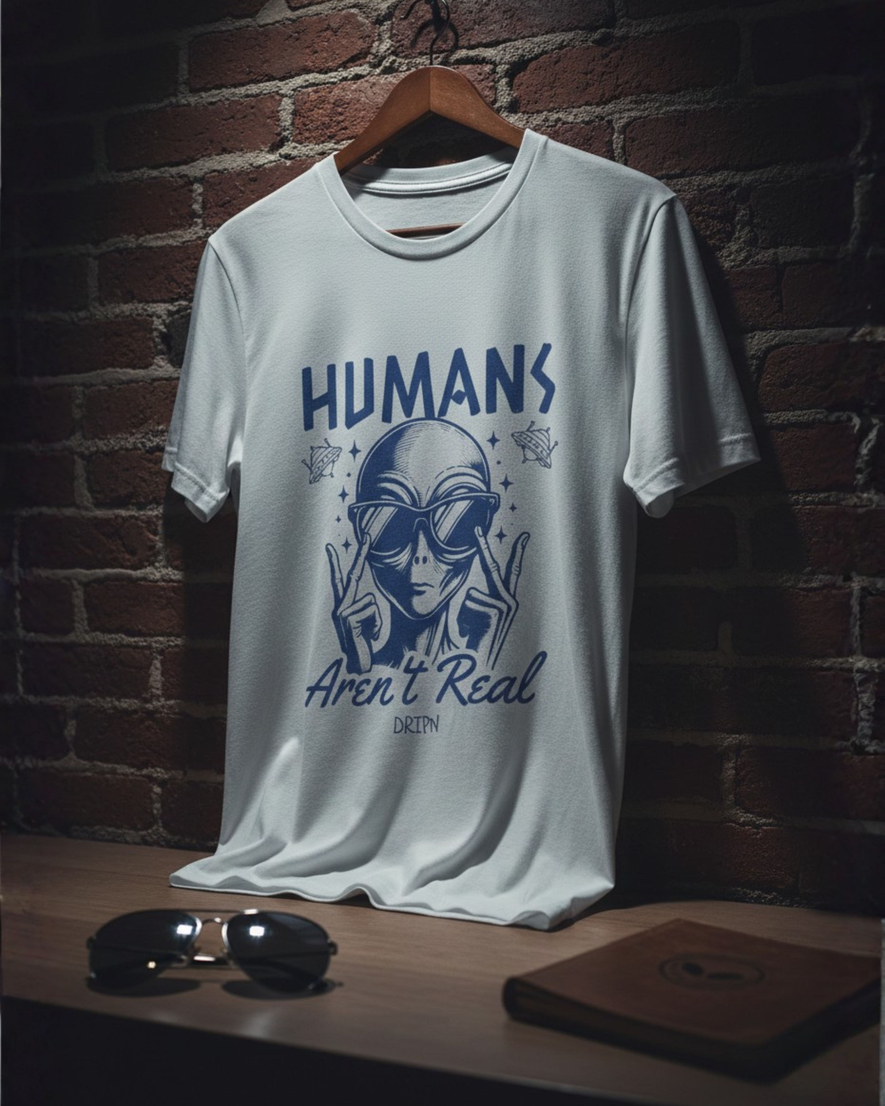 White t-shirt with alien graphic and text 'Humans Aren't Real' on a wooden hanger against a brick wall.